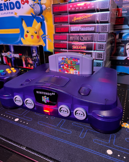 Front view of a Funtastic grape purple Nintendo 64 console with Super Mario 64 cartridge inserted and power light turned on.